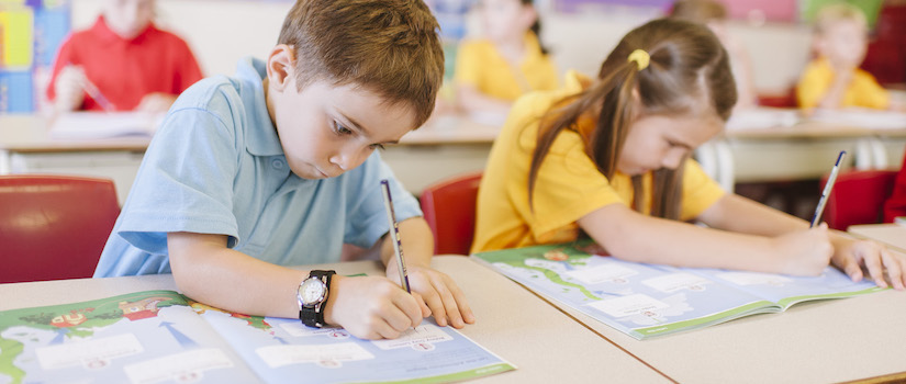 “students-using-writing-time-book-at-desk-in-classroom“